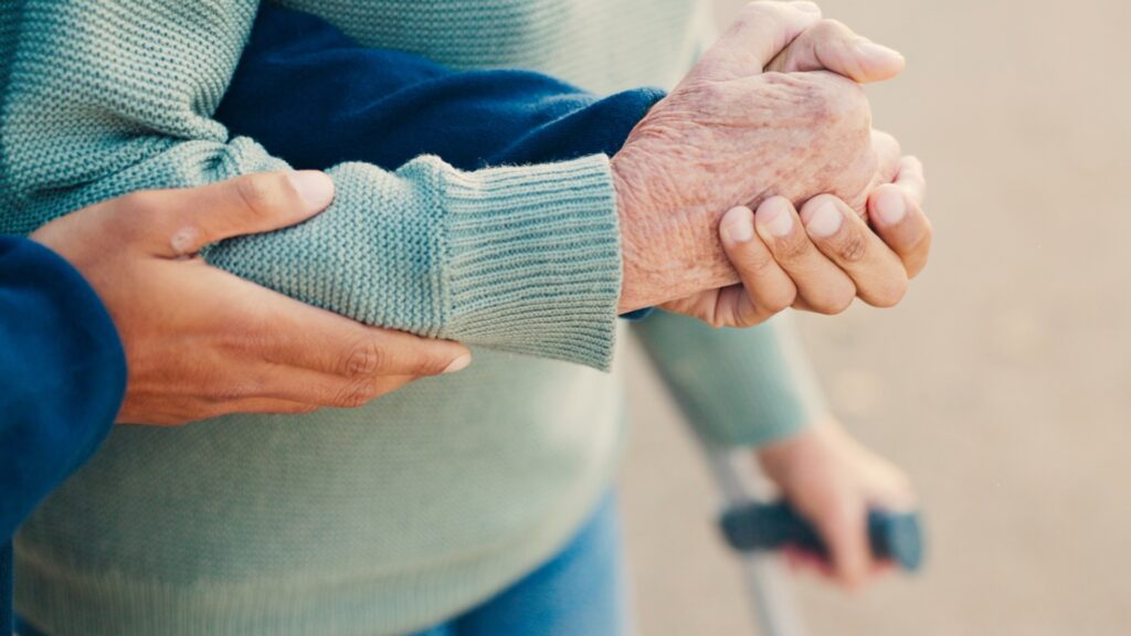 eine Hand einer Person hält eine andere ältere Hand, die in einer anderen Hand einen Gehstock hält