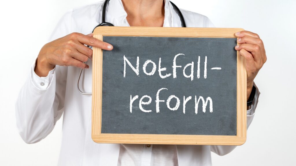 Mensch mit weißem Kittel hält Tafel mir der Aufschrift Notfallreform und zeigt mit dem Finger drauf.