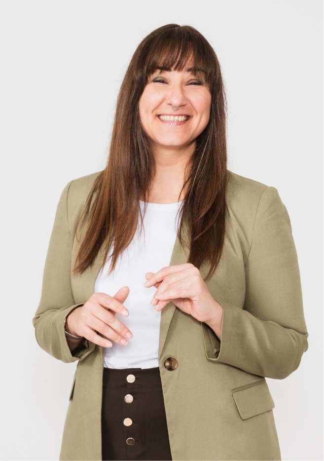 Frau mit braunen langen Haaren, einem, grünen Blazer und einem weißen Tshirt, schwarze Hose mit Knopfleiste schaut grinsend in die Kamera