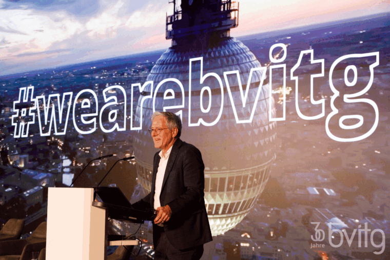 Matthias Meierhofer steht auf der Bühne am Pult und begrüßt das Publikum. Im Hintergrund sieht man den Fernsehrturm auf einer Leinwand mit der aufschrift #wearebvitg